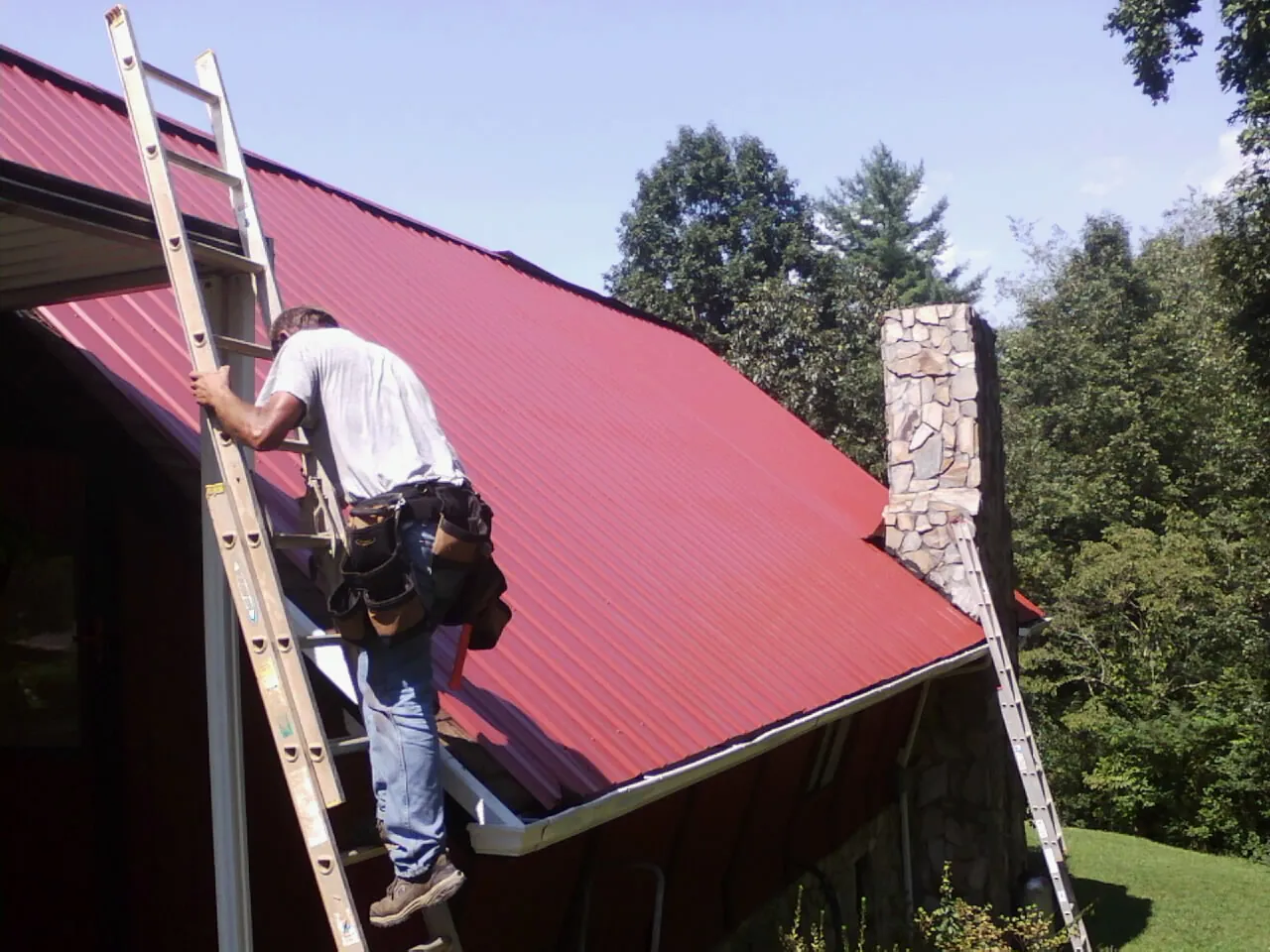 Roofer on a ladder beside a red metal roof and stone chimney — Soffit & Fascia Repair by Roofing Brothers in Fort Madison